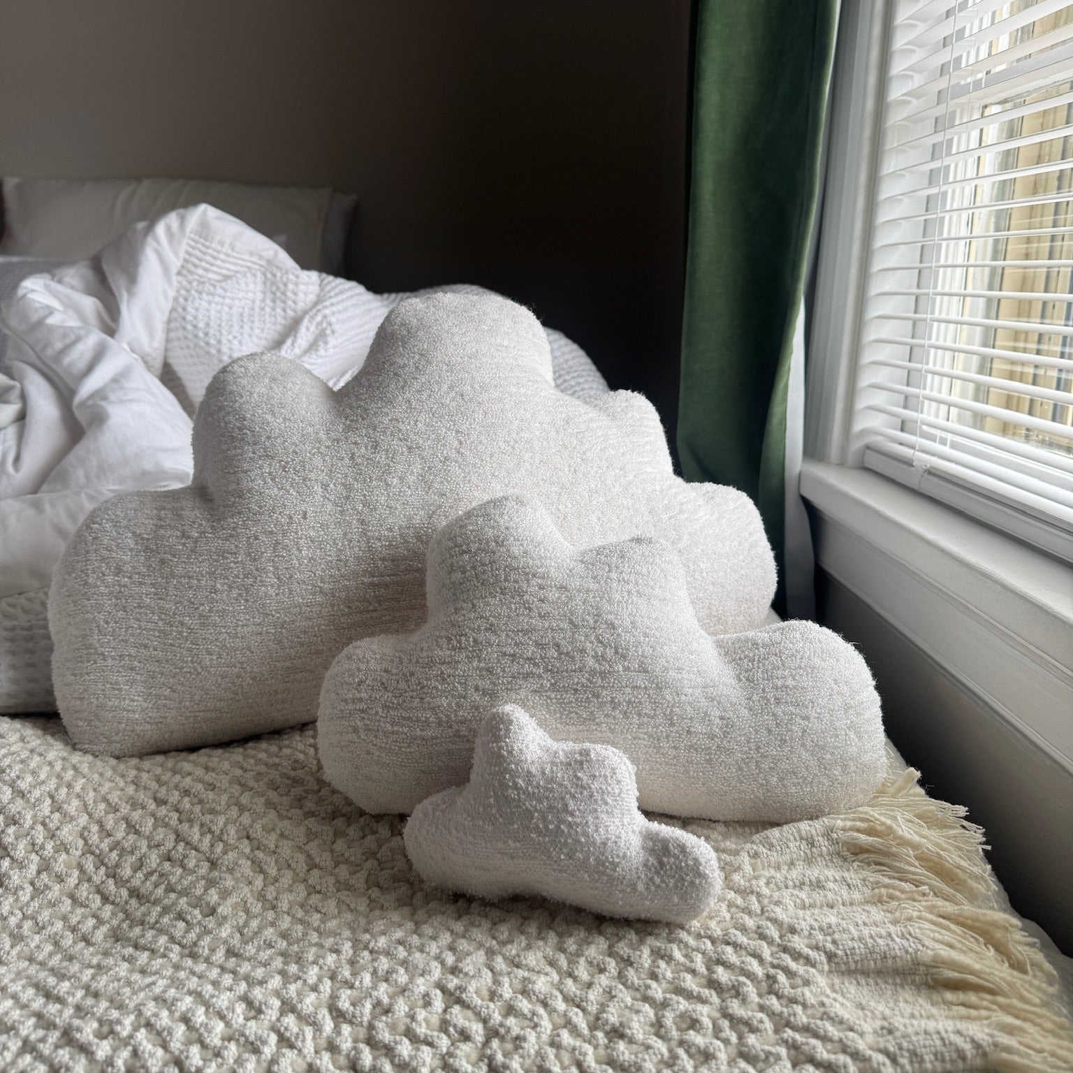 Cloud-shaped pillows on a bed next to a window with blinds and green curtains.