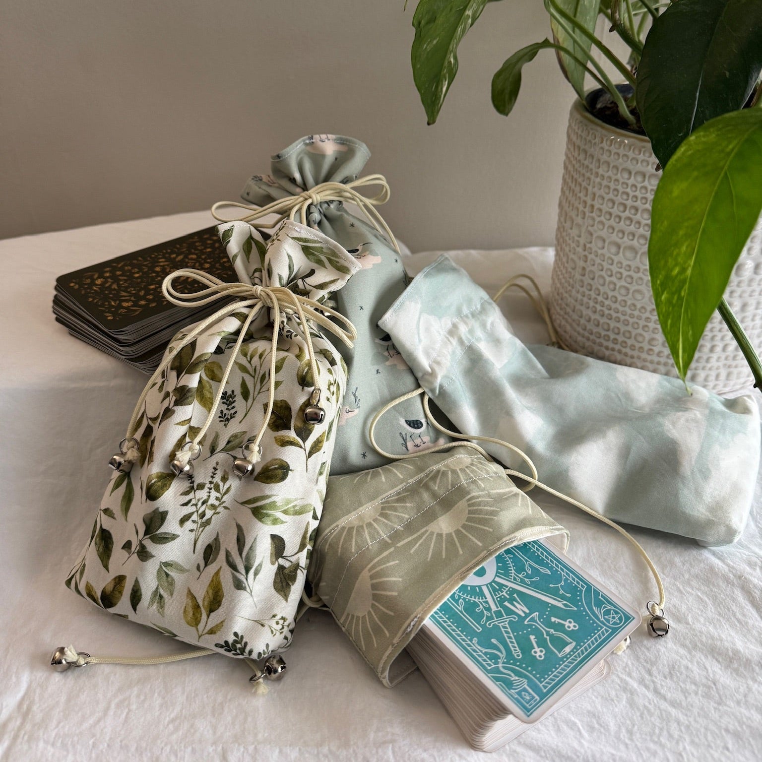 Drawstring bags with cool-toned designs on a white surface with a plant and tarot decks in the background.
