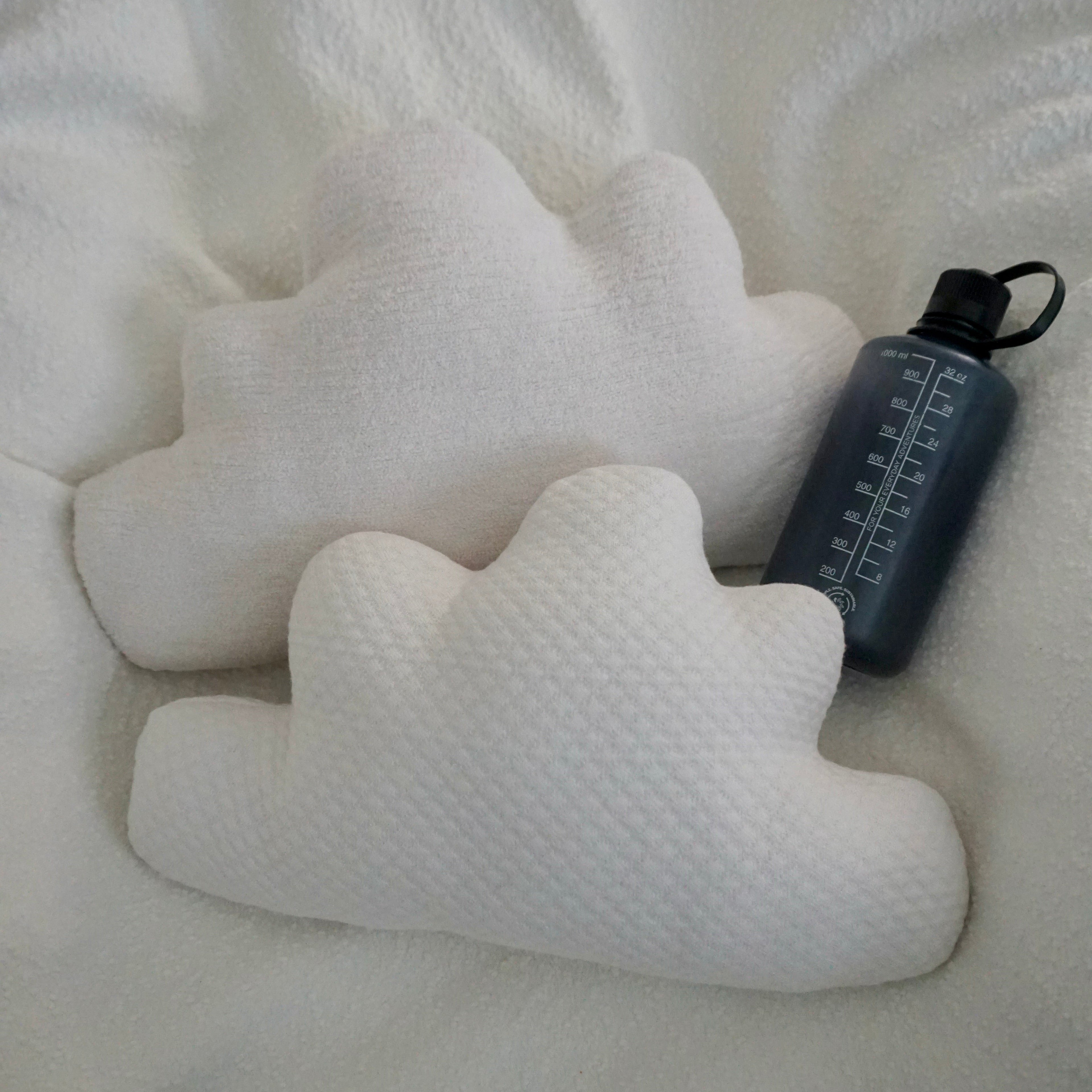 Two large Cloud Pillows arranged together on a textured white blanket with a water bottle for scale.