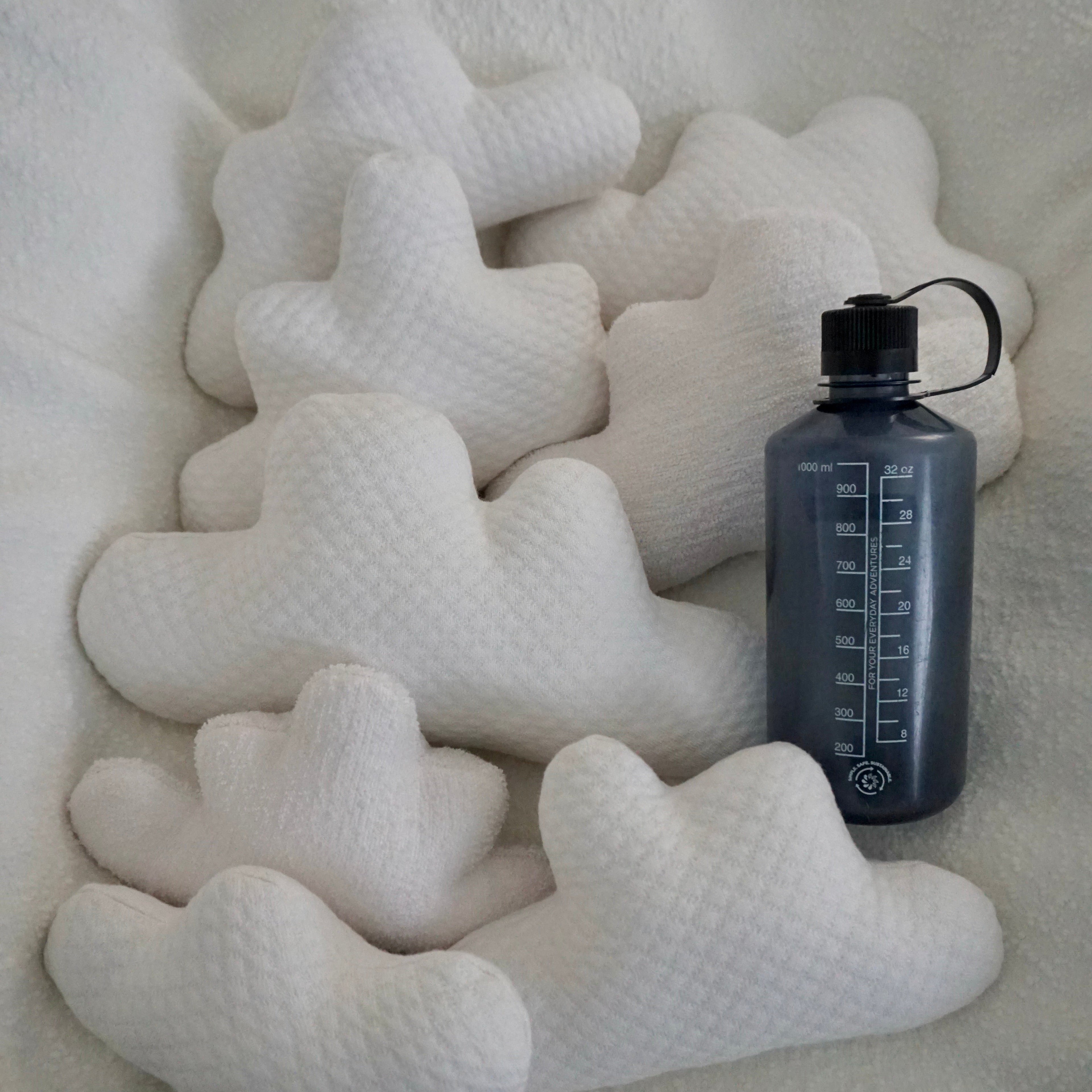 Eight medium Cloud Pillows arranged together on a textured white blanket with a water bottle for scale.