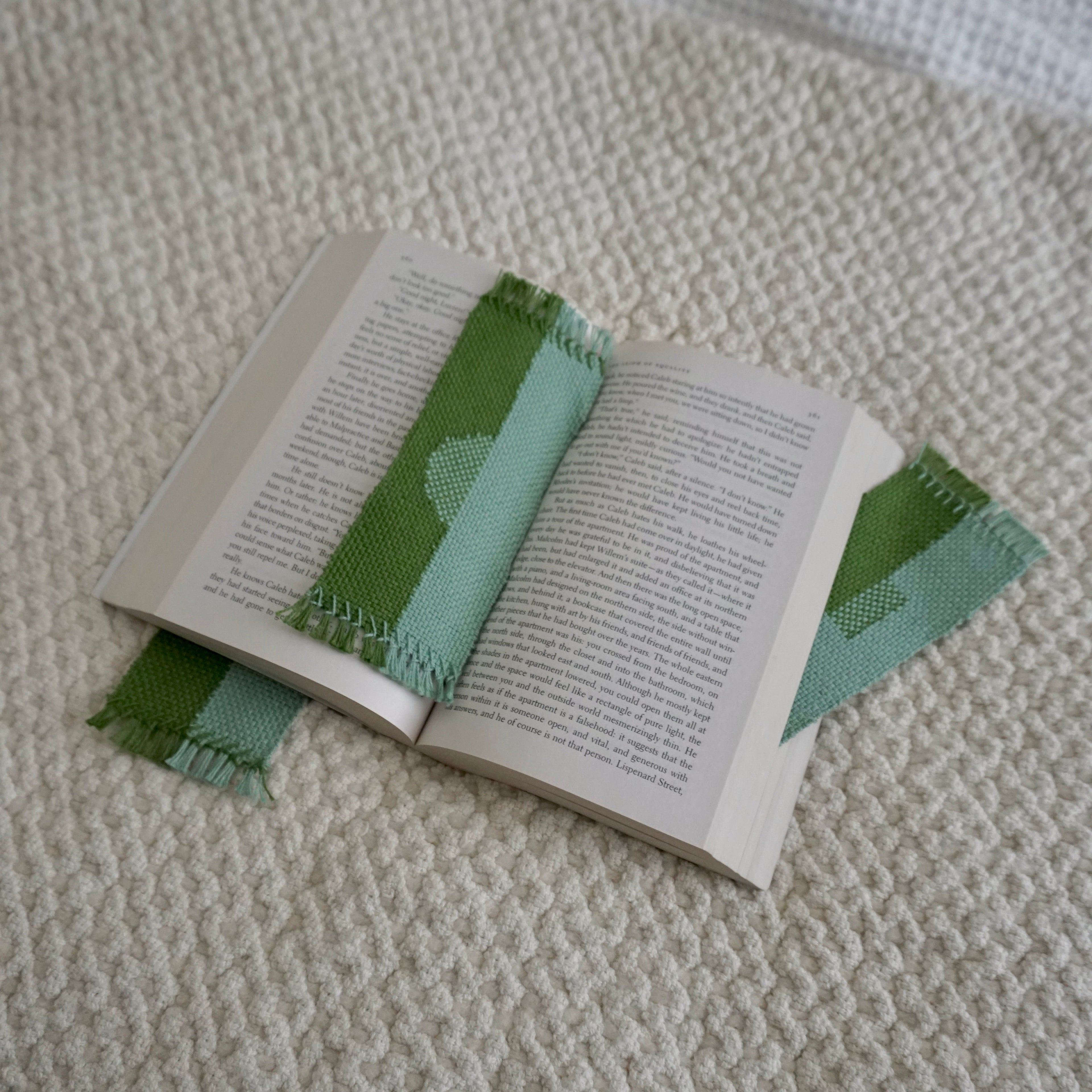 Three green and blue woven bookmarks inside an open book on a beige textured blanket.