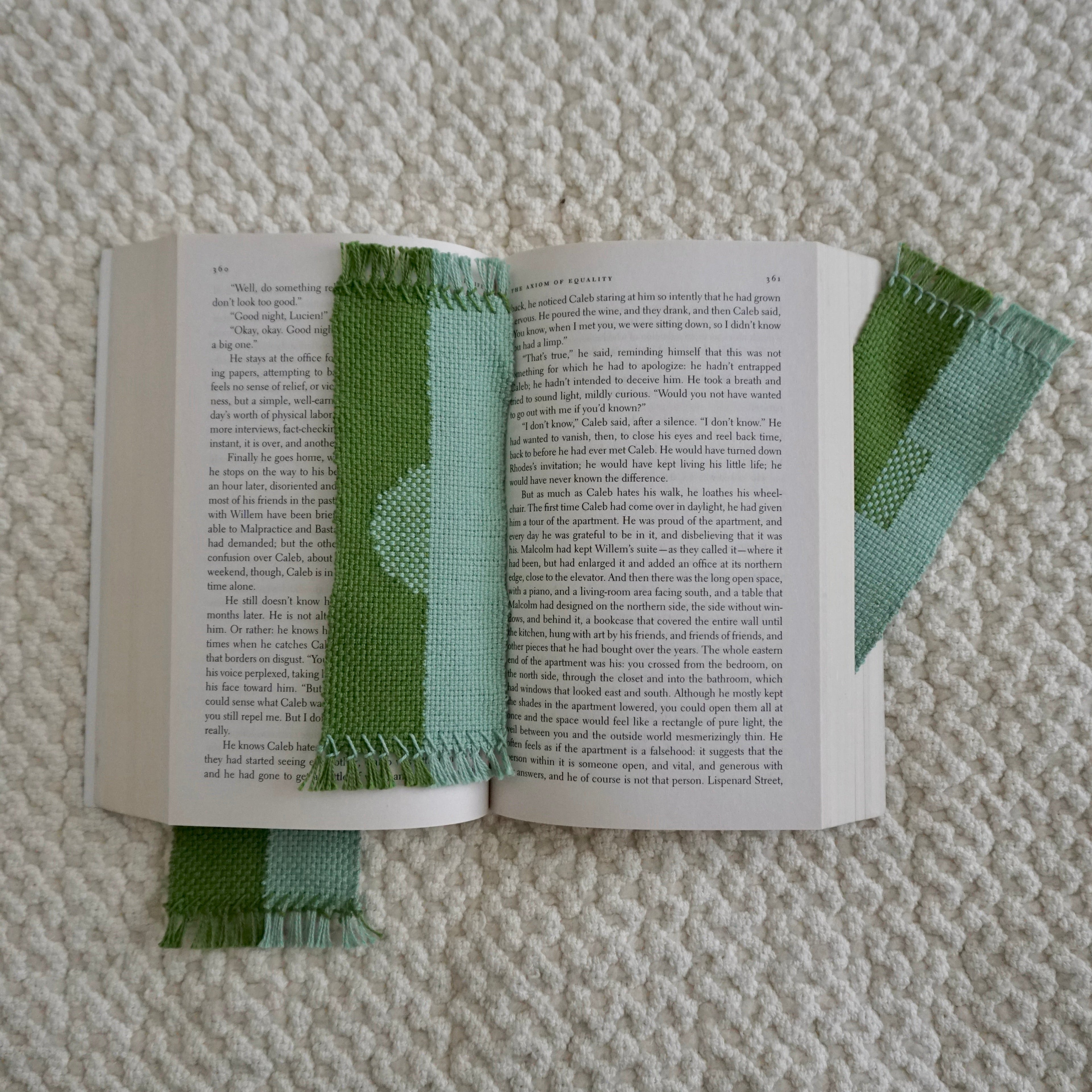 Three green and blue woven bookmarks in an open book resting on a textured woven blanket.